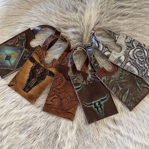 May include: A collection of handcrafted leather wine bottle tags. Each tag features unique embossed designs, including turquoise accents, a bull skull, and floral patterns. The tags have a loop for easy hanging.