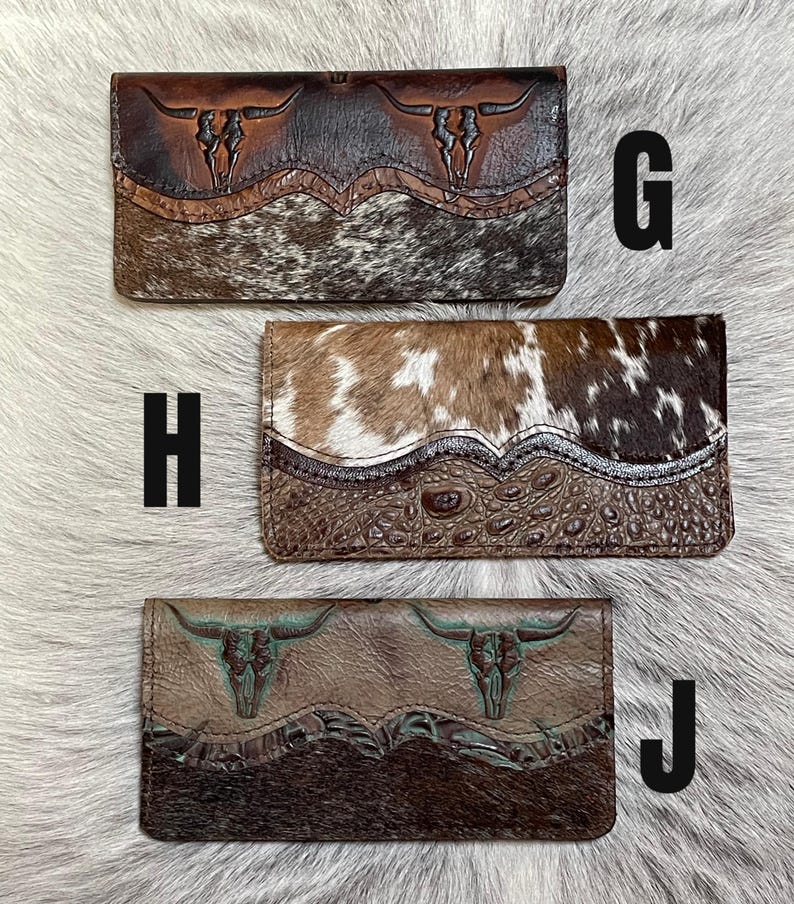 May include: Three rectangular leather wallets labeled G, H, and J. Wallet G features brown embossed steer skulls and cowhide. Wallet H has a brown and white cowhide top with an embossed crocodile texture bottom. Wallet J shows steer skulls and dark fur.