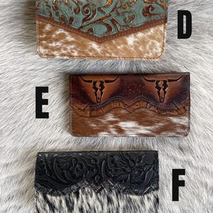 May include: Three rectangular wallets labeled D, E, and F. Wallet D has a teal floral embossed flap over brown cowhide. Wallet E features a dark brown leather flap with two embossed bull skulls over brown cowhide. Wallet F has a black floral embossed flap over black and white cowhide.