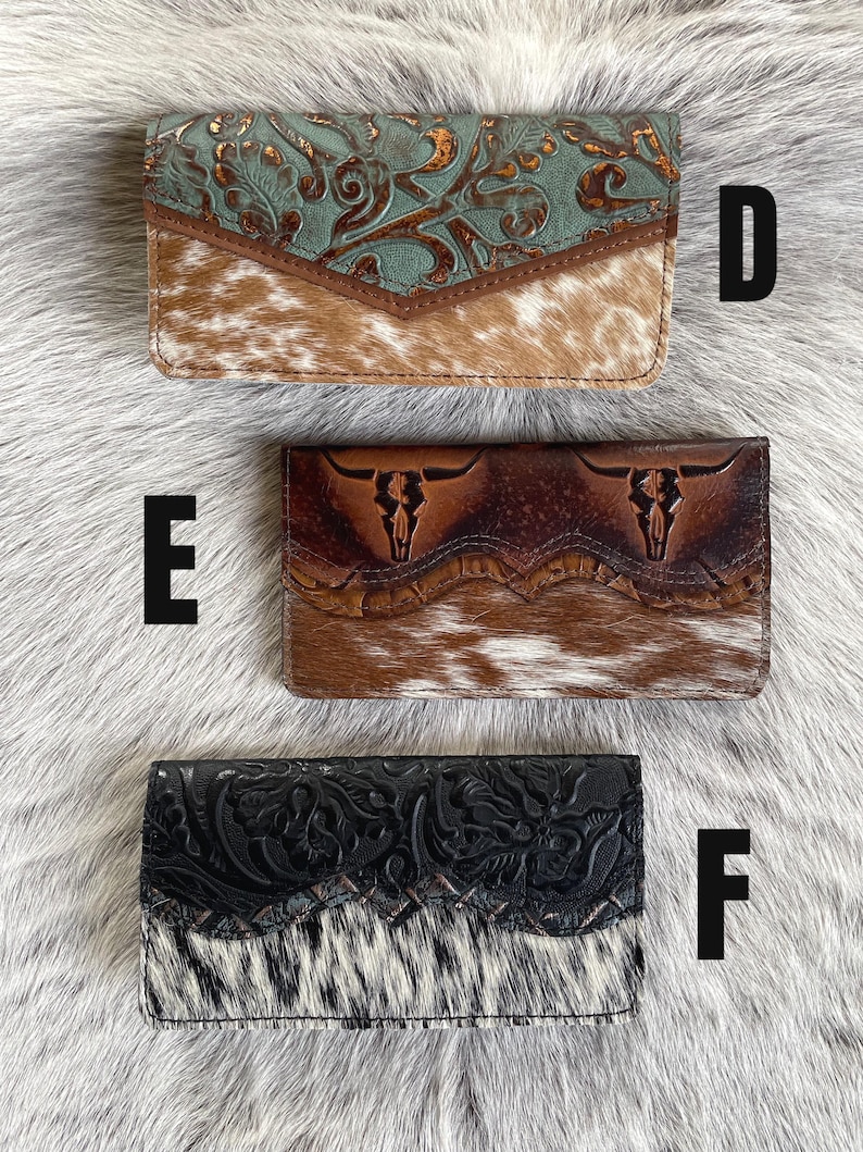 May include: Three rectangular wallets labeled D, E, and F. Wallet D has a teal floral embossed flap over brown cowhide. Wallet E features a dark brown leather flap with two embossed bull skulls over brown cowhide. Wallet F has a black floral embossed flap over black and white cowhide.