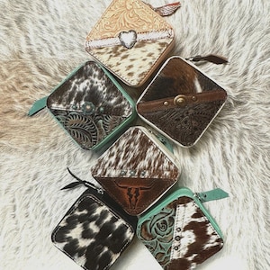 May include: A collection of square, zippered cowhide wallets with various designs. Some feature embossed leather, a heart accent, or a steer skull. The wallets have teal or brown trim and a leather pull tab.
