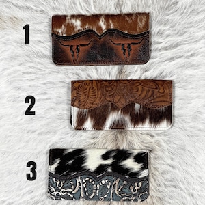 May include: Three rectangular wallets labeled 1, 2, and 3. Wallet 1 features brown and white cowhide with a dark brown embossed steer skull design. Wallet 2 has a brown floral embossed leather flap over brown and white cowhide. Wallet 3 has black and white cowhide with a blue floral embossed leather base.