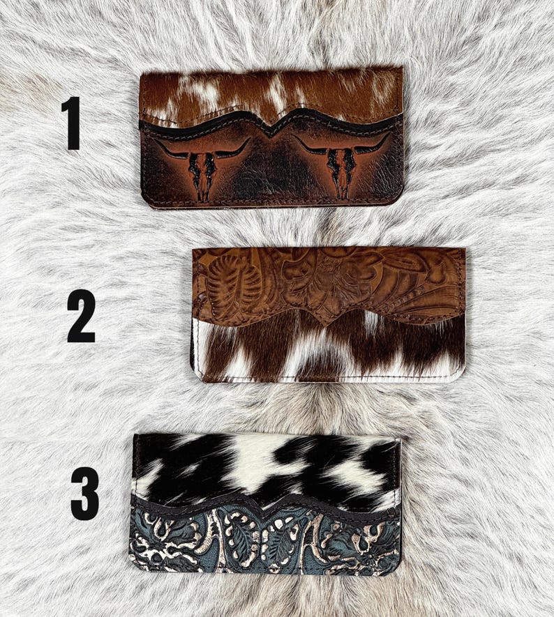 May include: Three rectangular wallets labeled 1, 2, and 3. Wallet 1 features brown and white cowhide with a dark brown embossed steer skull design. Wallet 2 has a brown floral embossed leather flap over brown and white cowhide. Wallet 3 has black and white cowhide with a blue floral embossed leather base.
