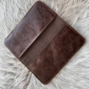 May include: A dark brown leather checkbook cover featuring a smooth, distressed finish on the outer panels and a textured, pebbled grain leather section in the center. The rectangular accessory has stitched edges and a slim, flat profile.