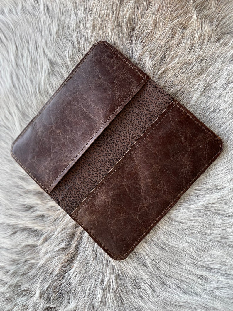 May include: A dark brown leather checkbook cover featuring a smooth, distressed finish on the outer panels and a textured, pebbled grain leather section in the center. The rectangular accessory has stitched edges and a slim, flat profile.