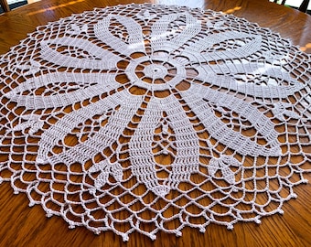 May include: A white crocheted table doily with a delicate floral design. The doily is circular and has a scalloped edge.