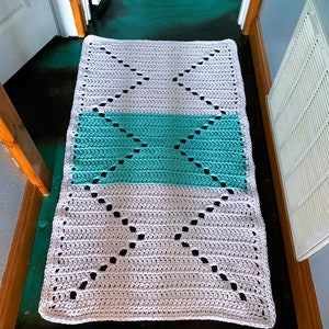 May include: A crocheted rug with a diamond pattern in white and teal. The rug is laid out on a dark green floor.