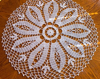 May include: White crocheted doily with a delicate floral design. The doily is circular and has a center with a smaller, more intricate design.