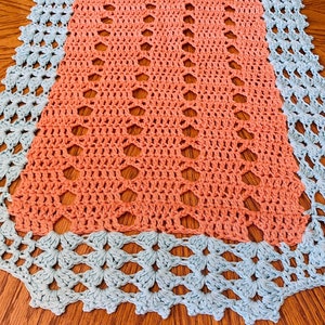 May include: A crocheted table runner with a peach colored center and a light blue border. The runner has a delicate, lacy pattern.