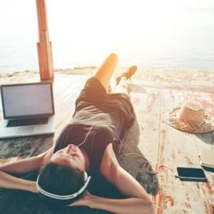 Peut inclure: Une femme portant un casque et une chemise noire est allongée sur une terrasse en bois avec un ordinateur portable, un chapeau de paille et un téléphone. La mer est visible en arrière-plan.