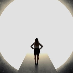 May include: Silhouette of a person standing on a platform, arms akimbo, facing a large, bright, circular opening. The person is wearing a white shirt and a black skirt. The background is a gradient of light.
