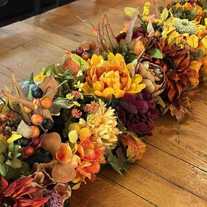 Puede incluir: Un camino de mesa con tem&aacute;tica oto&ntilde;al con una variedad de flores artificiales en tonos naranja, amarillo, rojo y marr&oacute;n. El camino de mesa incluye girasoles, crisantemos y otras hojas de oto&ntilde;o.