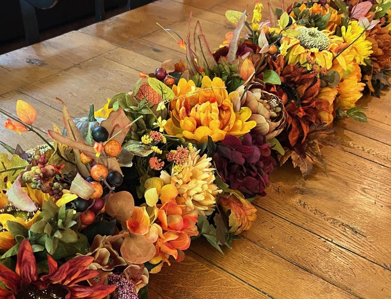Puede incluir: Un camino de mesa con tem&aacute;tica oto&ntilde;al con una variedad de flores artificiales en tonos naranja, amarillo, rojo y marr&oacute;n. El camino de mesa incluye girasoles, crisantemos y otras hojas de oto&ntilde;o.