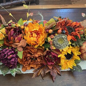 Puede incluir: Un centro de mesa de tem&aacute;tica oto&ntilde;al con una mezcla de flores naranja, amarillo, burdeos y marr&oacute;n. El centro de mesa incluye girasoles, peon&iacute;as y otras flores oto&ntilde;ales.