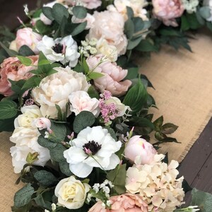 May include: A floral garland with white, pink, and cream flowers, including roses, peonies, and anemones. The garland is accented with greenery and small white flowers.