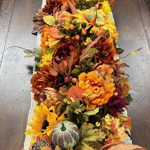 Puede incluir: Un camino de mesa con tem&aacute;tica oto&ntilde;al con una variedad de flores, hojas y calabazas artificiales. El camino de mesa presenta girasoles, peon&iacute;as naranja y burdeos y una peque&ntilde;a calabaza verde.