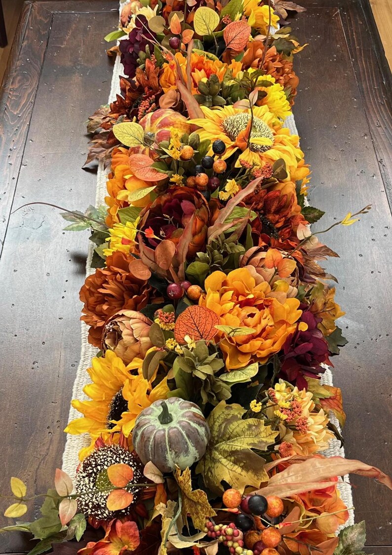 Puede incluir: Un camino de mesa con tem&aacute;tica oto&ntilde;al con una variedad de flores, hojas y calabazas artificiales. El camino de mesa presenta girasoles, peon&iacute;as naranja y burdeos y una peque&ntilde;a calabaza verde.