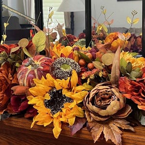 Puede incluir: Un centro de mesa de oto&ntilde;o con girasoles, peon&iacute;as burdeos y naranja, una peque&ntilde;a calabaza y follaje oto&ntilde;al.