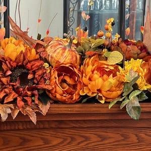 Puede incluir: Un arreglo floral de oto&ntilde;o con flores naranja, amarillas y marrones, hojas y una peque&ntilde;a calabaza naranja. El arreglo se muestra sobre una superficie de madera.
