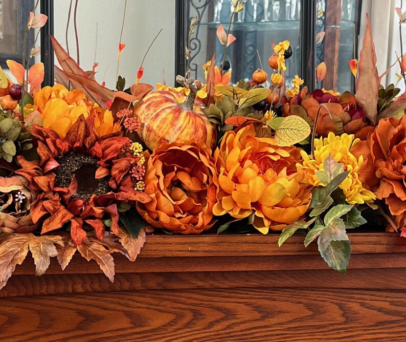 Puede incluir: Un arreglo floral de oto&ntilde;o con flores naranja, amarillas y marrones, hojas y una peque&ntilde;a calabaza naranja. El arreglo se muestra sobre una superficie de madera.