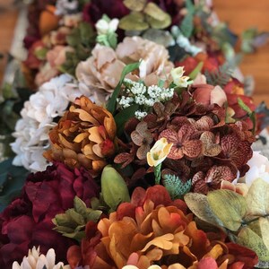 May include: A close-up of a floral arrangement featuring a variety of artificial flowers in shades of burgundy, orange, cream, and green. The arrangement includes peonies, hydrangeas, and other decorative blooms, creating a rich, textured display.