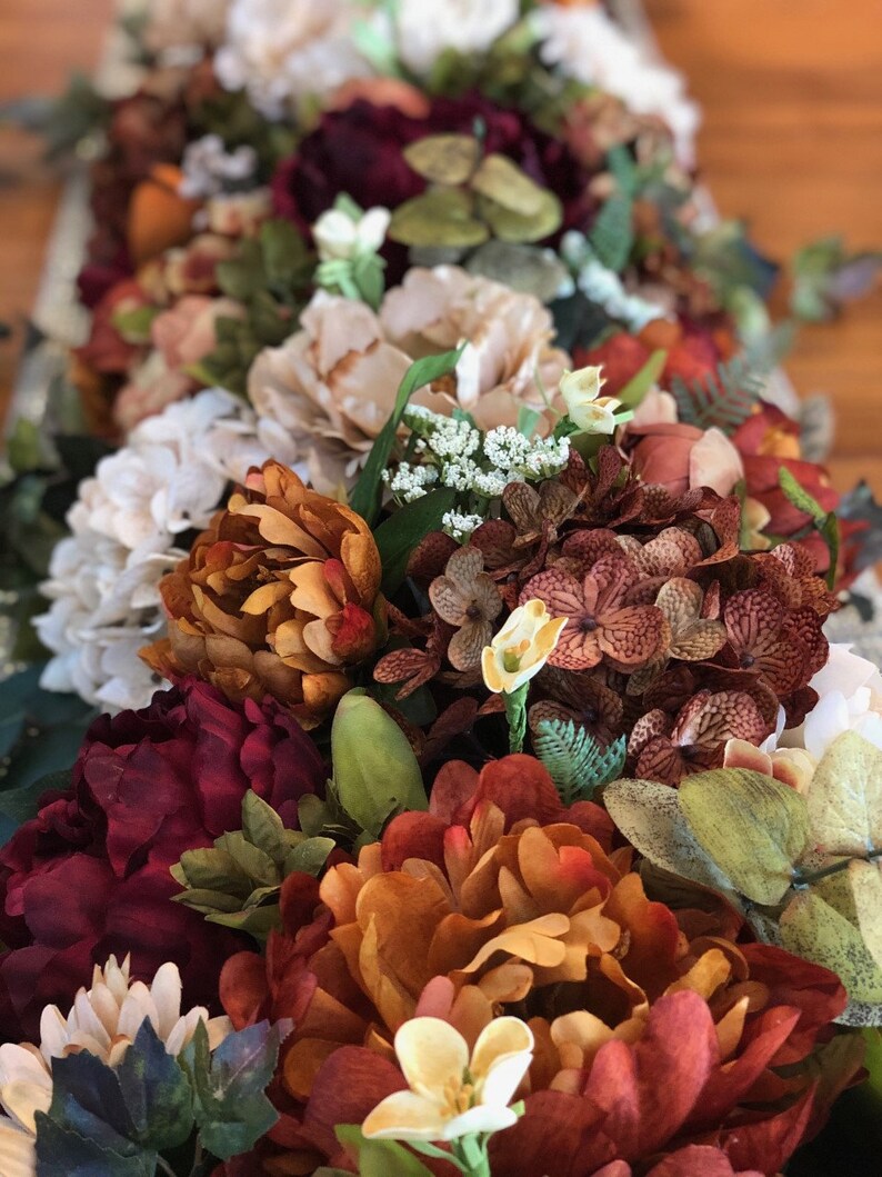 May include: A close-up of a floral arrangement featuring a variety of artificial flowers in shades of burgundy, orange, cream, and green. The arrangement includes peonies, hydrangeas, and other decorative blooms, creating a rich, textured display.