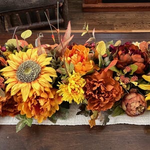Puede incluir: Un camino de mesa con tem&aacute;tica oto&ntilde;al con una variedad de flores artificiales, incluyendo girasoles, peon&iacute;as y crisantemos. Las flores son en tonos de amarillo, naranja y marr&oacute;n.