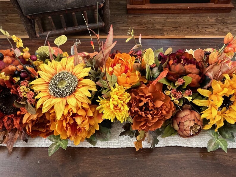 Puede incluir: Un camino de mesa con tem&aacute;tica oto&ntilde;al con una variedad de flores artificiales, incluyendo girasoles, peon&iacute;as y crisantemos. Las flores son en tonos de amarillo, naranja y marr&oacute;n.