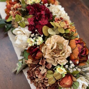May include: A floral arrangement featuring a variety of flowers in shades of cream, burgundy, orange, and brown. The arrangement includes peonies, hydrangeas, and other flowers, with green foliage. The flowers are arranged on a cream-colored fabric.