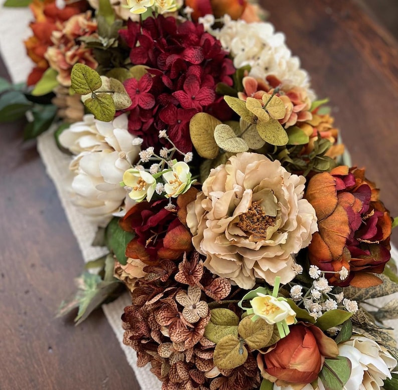 May include: A floral arrangement featuring a variety of flowers in shades of cream, burgundy, orange, and brown. The arrangement includes peonies, hydrangeas, and other flowers, with green foliage. The flowers are arranged on a cream-colored fabric.
