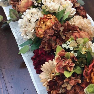 May include: A floral arrangement featuring a variety of artificial flowers in shades of cream, orange, burgundy, and brown. The arrangement includes peonies, hydrangeas, and other blooms, resting on a white runner with a subtle pattern. The overall theme is autumnal.