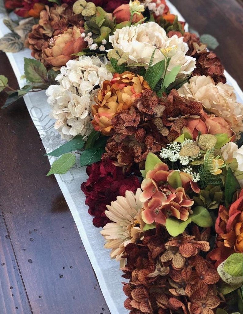 May include: A floral arrangement featuring a variety of artificial flowers in shades of cream, orange, burgundy, and brown. The arrangement includes peonies, hydrangeas, and other blooms, resting on a white runner with a subtle pattern. The overall theme is autumnal.