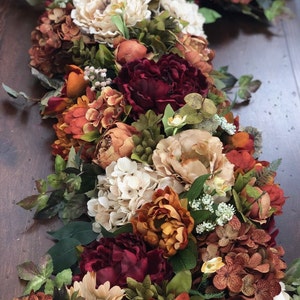 May include: A vibrant floral arrangement featuring a variety of faux flowers in warm autumn colors. The arrangement includes peonies, hydrangeas, and other blooms in shades of burgundy, orange, and cream, with green foliage accents. Perfect for fall decor.