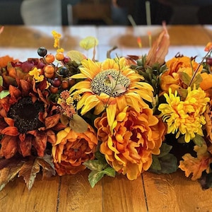 Puede incluir: Un centro de mesa floral de tem&aacute;tica oto&ntilde;al con girasoles, peon&iacute;as y otras flores oto&ntilde;ales en tonos naranja, amarillo y burdeos. El centro de mesa est&aacute; dispuesto sobre una mesa de madera.