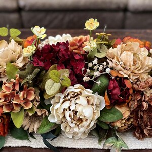 May include: A vibrant floral arrangement featuring a mix of faux flowers in shades of cream, burgundy, orange, and yellow. The arrangement includes peonies, hydrangeas, and other greenery, creating a full and textured display. Perfect for home decor.