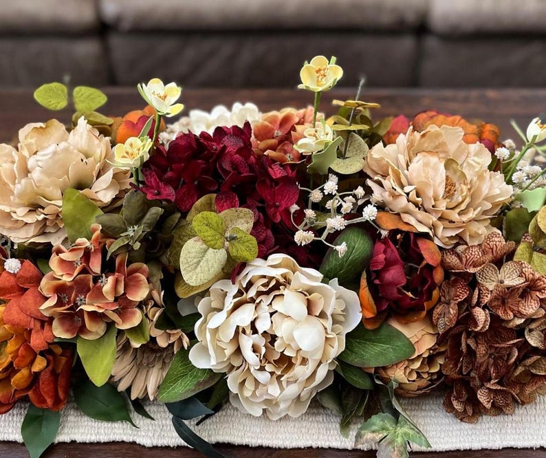 May include: A vibrant floral arrangement featuring a mix of faux flowers in shades of cream, burgundy, orange, and yellow. The arrangement includes peonies, hydrangeas, and other greenery, creating a full and textured display. Perfect for home decor.