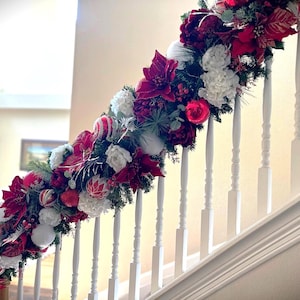 Puede incluir: Un pasamanos blanco decorado con una guirnalda navideña roja y blanca. La guirnalda presenta flores de poinsettia, bolas blancas y adornos rojos.