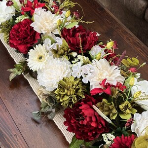 May include: A long, rustic table runner with a burlap base is adorned with a lush arrangement of red and white flowers. The centerpiece features white peonies, red peonies, white gerbera daisies, and green foliage.