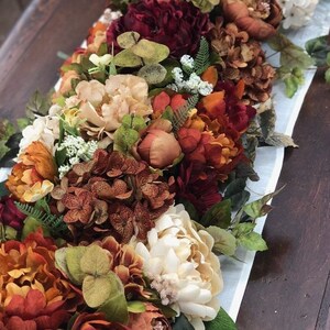 May include: A floral table runner featuring an arrangement of faux flowers in shades of orange, red, cream, and green. The runner includes peonies, hydrangeas, and other decorative blooms, set on a light blue and white striped fabric.