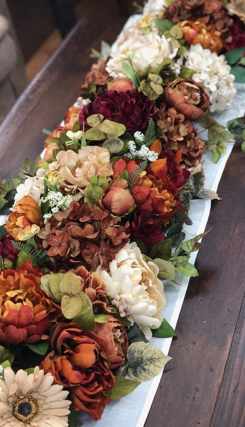 May include: A floral table runner featuring an arrangement of faux flowers in shades of orange, red, cream, and green. The runner includes peonies, hydrangeas, and other decorative blooms, set on a light blue and white striped fabric.