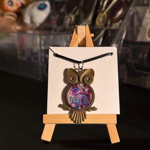 May include: A bronze-colored owl pendant necklace. The pendant features a circular design with the text "TAPE B" and a colorful, abstract image. The necklace is displayed on a small wooden easel.