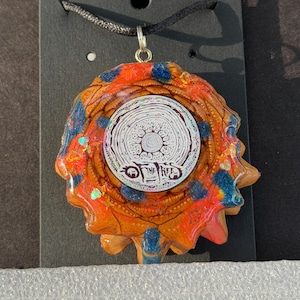 May include: A circular pendant featuring a central white disc with black intricate line art and the text OF THE TREES. The surrounding border is a textured, resin-coated pinecone segment with vibrant orange, red, and blue glitter accents.