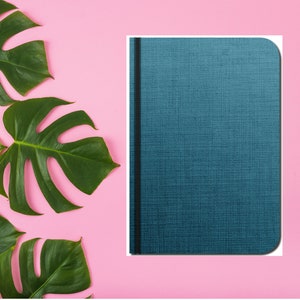 Puede incluir: Un cuaderno azul con una encuadernaci&oacute;n negra y una cubierta texturizada. El cuaderno est&aacute; sobre un fondo rosa con hojas verdes.