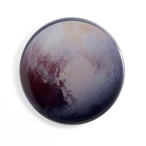 May include: A close-up of a round button featuring a colorful image of Pluto, a dwarf planet in our solar system. The image shows a swirling mix of purple, blue, and brown hues.