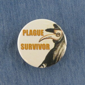 May include: A white button with a black and white illustration of a plague doctor wearing a mask and hat. The text "PLAGUE SURVIVOR" is printed in orange on the button.