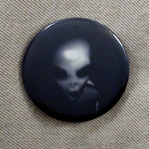 May include: A black and white button with a blurry image of a grey alien's face.