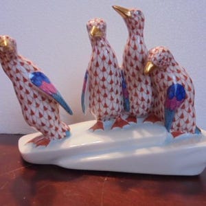 May include: Four porcelain penguins with red, white, and blue patterned bodies stand on a white base. The penguins are arranged in a row, with the two on the ends facing forward and the two in the middle facing each other.