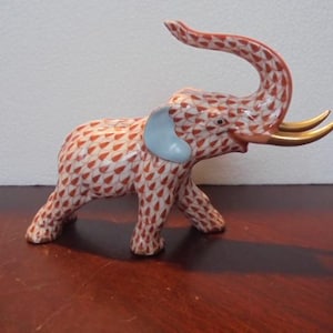 May include: A red and white porcelain elephant figurine with a raised trunk and gold-colored tusks. The elephant is standing on all four legs and has a blue ear.