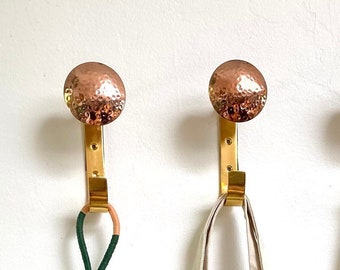 May include: Three gold hooks with hammered copper finials hang on a white wall. A white canvas tote bag with black lettering that reads "AD ARCHITECTURAL DIGEST" hangs on one hook. A green and pink beaded chain with small bells hangs on another hook.