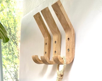 May include: Three wooden wall hooks with a curved design are mounted on a white wall. A hanging plant in a woven basket is suspended below the hooks. The plant has green and white leaves.